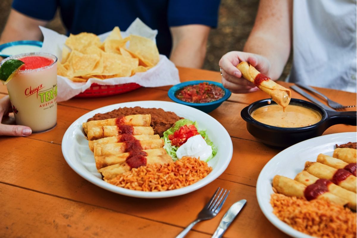Three people sitting at a bench and sharing two plates of crispy flautas.