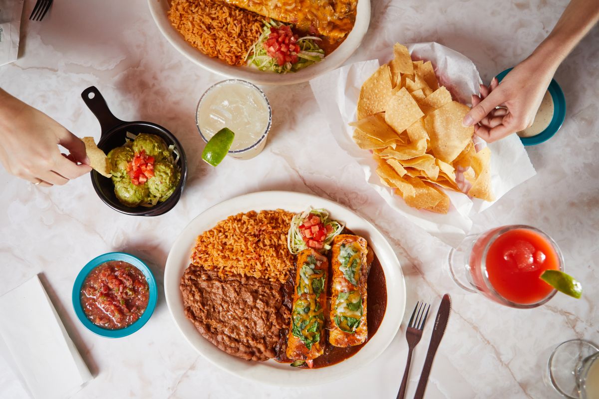 Overhead shot of two hands on a marble table with enchiladas, chips, guac and margaritas.