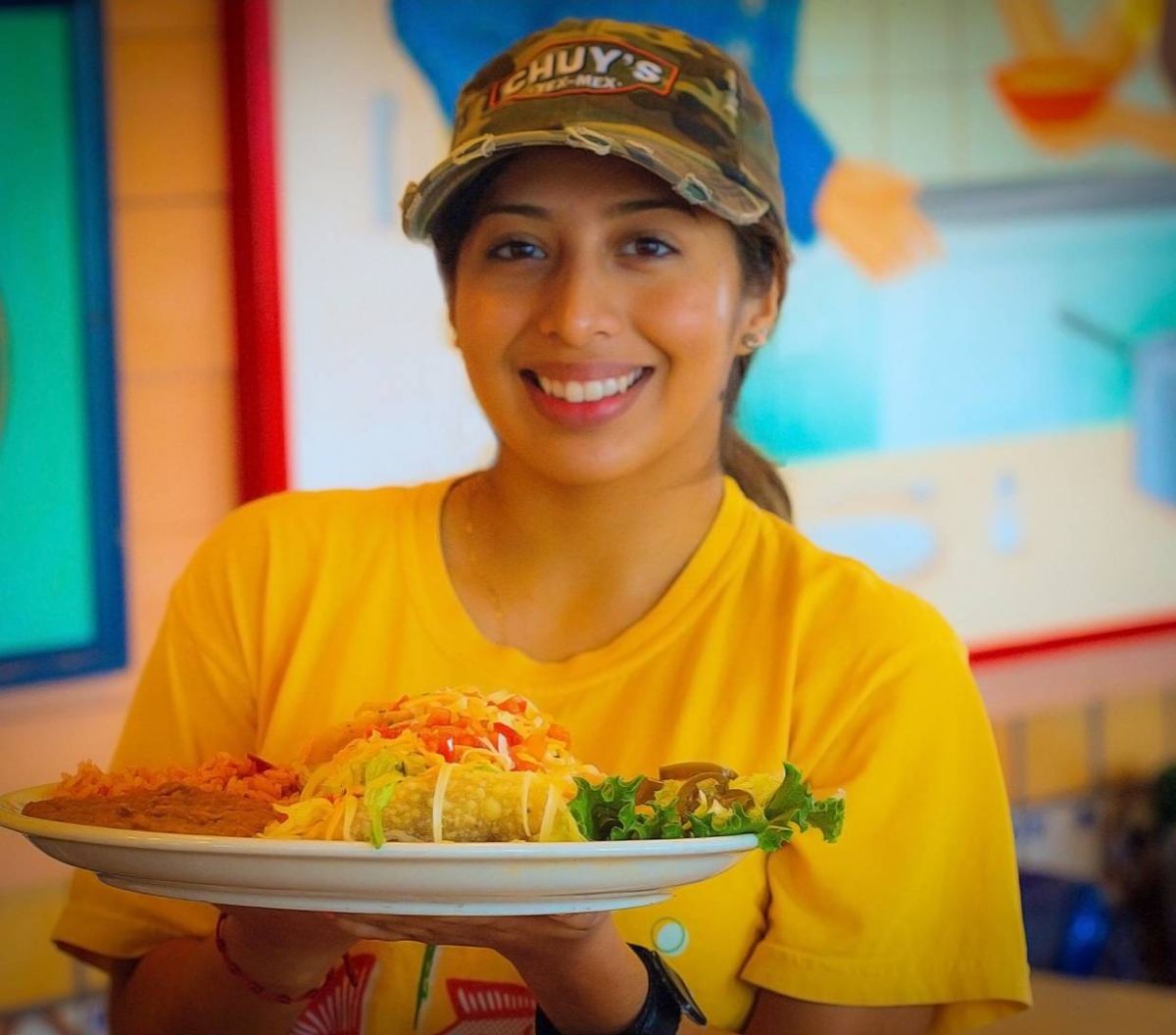 A Chuy’s employee holds a plate of tacos while wearing a Chuy’s camo hat and a yellow t-shirt.